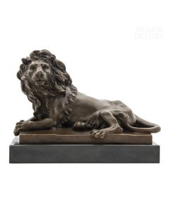 Dekor Deluxe kovinska figura sproščenega leva bronaste barve, ki leži na črnem piedestalu. Visoka 20 cm in široka 27 cm.