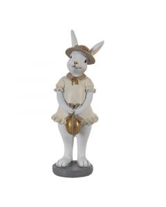 Figura belega velikonočnega zajčka zajčice v bež krilu s rjavim klobukom in z zlatim jajcem v rokah. 